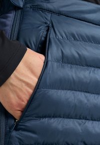 Marineblauwe gewatteerde jas met een horizontaal doorgestikt patroon. Zichtbare hand in de met rits afgesloten zak. Glad, lichtgewicht stof.