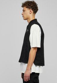 Melnā veste ar augstu apkakli un rāvējslēdzēju, ar divām priekšējām kabatām. Valkāta virs baltas īsām piedurknēm krekla ar gludu auduma tekstūru.