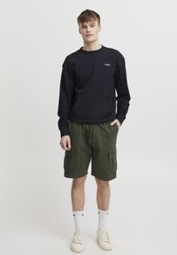 Svart hettegenser med frontlomme og logo, sammen med grønne cargo-shorts med sidelommer. Modellen har på seg hvite sokker og sneakers.