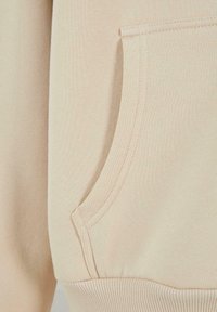 Tessuto di felpa beige con una consistenza morbida, dotato di una tasca laterale e polsini a coste. Le cuciture lungo la tasca evidenziano il suo design.