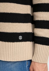 Gestreifter Pullover in Schwarz und Beige, strukturiertes Strickdesign, gerippte Bündchen und Saum, mit einem kleinen OXMO-Logo an der Unterseite.