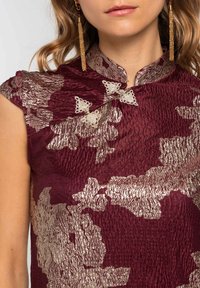 Vestido de seda burdeos con patrones florales texturizados en hilo dorado. Presenta un cuello mandarín y cierres decorativos, exhibiendo detalles intrincados.
