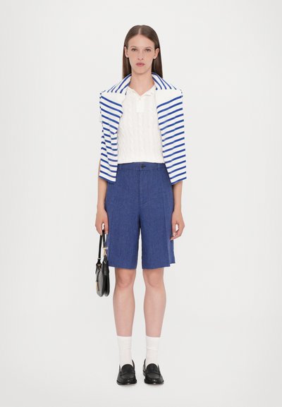 Polo en tricot blanc sous un blazer rayé bleu et blanc, associé à un short bleu. Des mocassins noirs et un petit sac à main noir complètent le look.