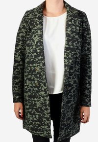 Abrigo de patrón negro y verde con cuello de solapa, mangas largas y dos botones en la parte delantera. Llevado sobre una blusa blanca y pantalones negros.