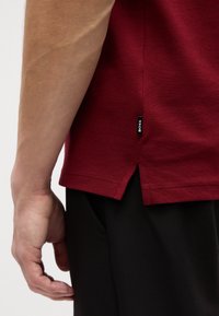 Burgundi polo marškiniai su tekstūruota medžiaga, šoninėmis vėdinimo detalėmis ir mažu juodu prekės ženklo etikete hemu, derinami su juodomis kelnėmis.