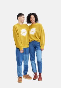 Des pulls en laine jaune au look surdimensionné avec un logo "Kickers" blanc. À associer avec un jean en patchwork bleu et des chaussures à lacets marron.