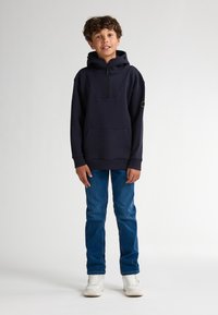 Felpa con zip e cappuccio di colore blu navy, con tasca a marsupio, polsini a coste e una toppa con logo sulla manica, abbinata a jeans azzurri e sneakers.