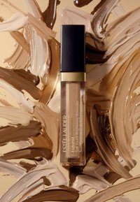 Estee Lauder Futurist concealer i en klar tube med gull- og svart hette, plassert på en bakgrunn av blandede hudtoner i kremfarge og brunt.