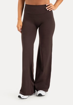 Femme portant un pantalon large taille haute marron foncé avec des baskets blanches, debout avec une main sur la hanche et l'autre détendue sur le côté.