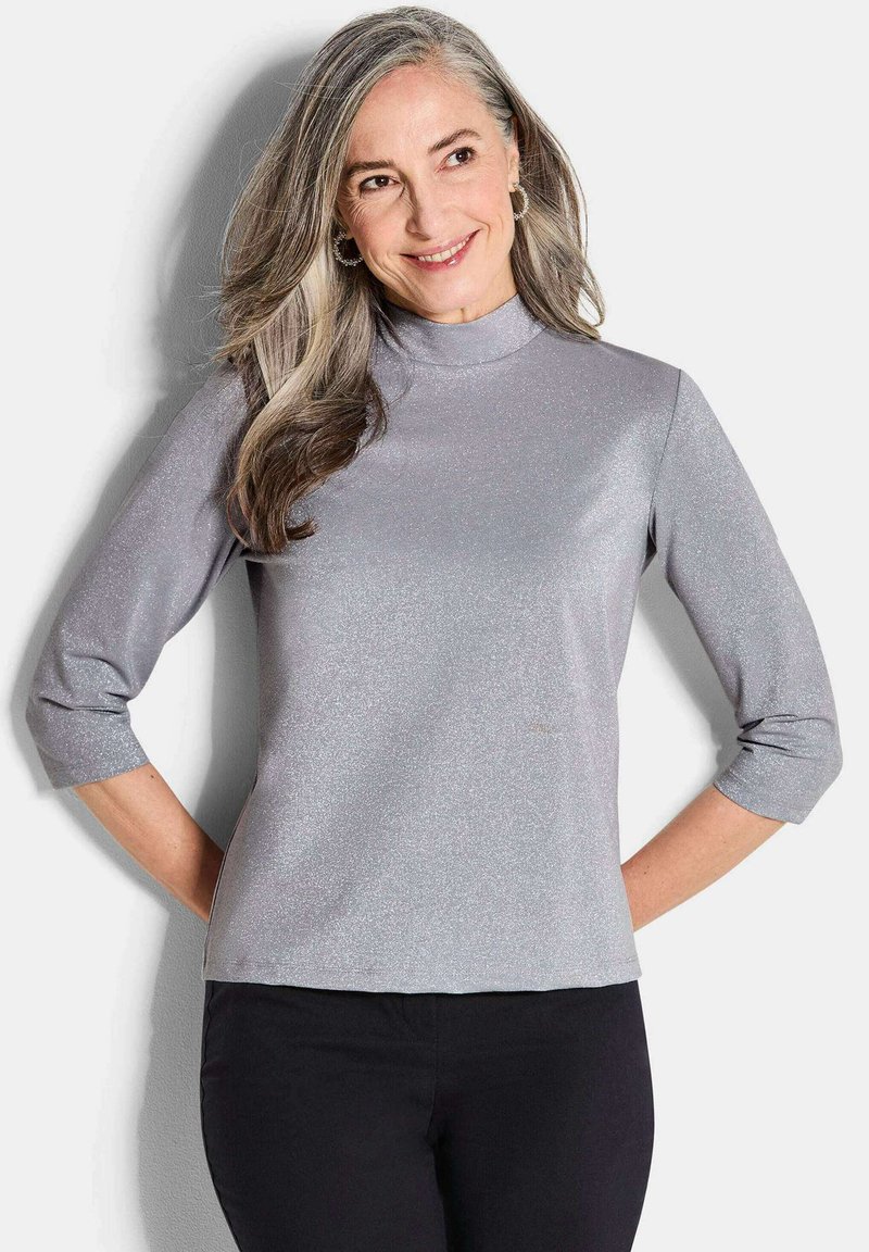 Maglietta argento a maniche lunghe con colletto alto e maniche a tre quarti, realizzata in un tessuto morbido e scintillante. Indossata con pantaloni neri.