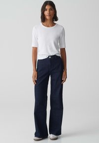 Top bianco a maniche corte abbinato a jeans a gamba larga blu scuro, con chiusura a bottone e un finish pulito. L'outfit è completato da sneakers chiare.