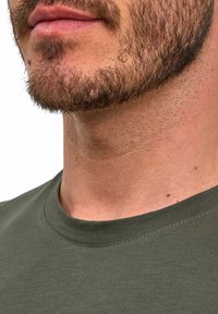 T-shirt in cotone verde oliva con scollo rotondo, che presenta una texture liscia e dettagli di cucitura minimi, con barba parzialmente visibile.
