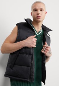 Gilet imbottito nero con colletto alto, dotato di chiusura con zip e tasche laterali, indossato sopra un maglione a maglia verde. Materiale testurizzato, design minimalista.