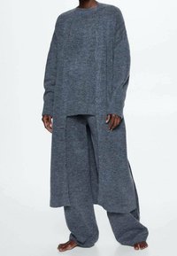 Szary, fantazyjnie dziergany, oversize'owy sweter z długimi rękawami i bocznymi rozcięciami, zestawiony z szerokimi spodniami o prostych nogawkach w odcieniu szarości. Miękka tekstura i luźne, swobodne dopasowanie na całej długości.