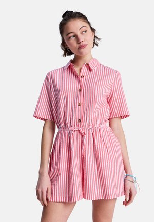 Młoda kobieta ubrana w różowo-białe krótkie kombinezon z pionowymi paskami, guzikami i paskiem w talii, patrząca w lewo.