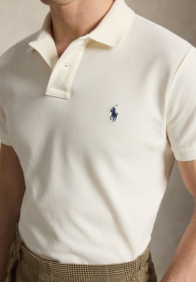 Mężczyzna ubrany w białą koszulkę polo z małym granatowym logo konia i jeźdźca, w parze z brązowymi spodniami w kratę.