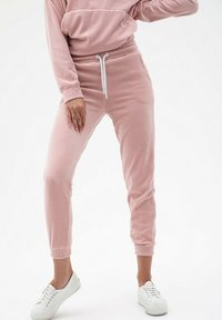 Różowe welurowe dresy z elastycznymi mankietami i paskiem w talii, z białym sznurkiem. W zestawieniu z białymi sneakersami na jednolitym tle.