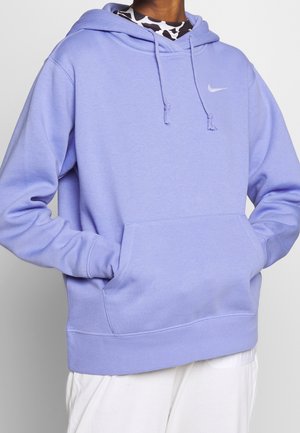 Osoba nosząca jasnofioletową bluzę z kapturem Nike z rękami w przedniej kieszeni i białe spodnie, stojąca na jednolitym tle.