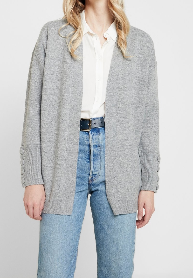 Femme portant un cardigan gris clair ouvert sur le devant avec des détails de boutons sur les manches, par-dessus un chemisier blanc et un jean bleu taille haute avec une ceinture noire.