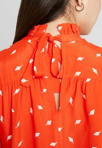 Rode blouse met een geschulpte halslijn, striksluiting en een sleutelgatdetail aan de achterkant, met een patroon van witte bladvormen op een gestructureerde stof.