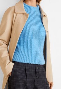 Kobieta w jasnobrązowym płaszczu, nałożonym na niebieski sweter o prążkowanej strukturze, oraz w ciemnych, kratkowanych spodniach o wysokim stanie z trzema guzikami.