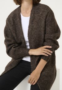 Brązowy, oversize sweter z dzianiny o miękkiej fakturze, z otwartym przodem i podwiniętymi rękawami, noszony na białej koszuli i czarnych spodniach.