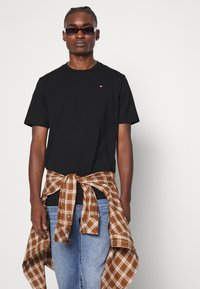 T-shirt nera in cotone con maniche corte, abbinata a una camicia a quadri marrone e crema legata in vita, indossata con jeans blu.