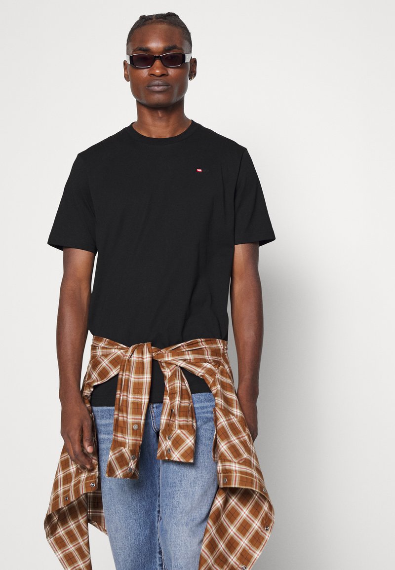 T-shirt nera in cotone con maniche corte, abbinata a una camicia a quadri marrone e crema legata in vita, indossata con jeans blu.