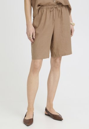 Person, die kniehohe braune Shorts mit Kordelzug, braune spitze Ballerinas, ein lockeres braunes Oberteil, einen Ring und ein Armband trägt und vor einem hellen Hintergrund steht.