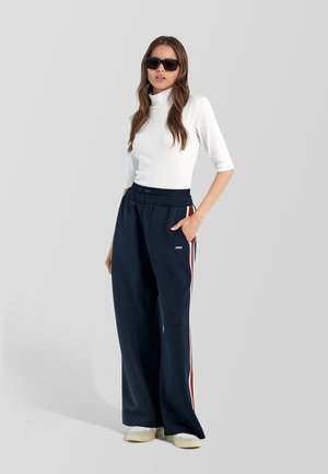 Mujer con gafas de sol negras, top blanco ajustado de cuello alto, pantalones anchos azul marino con rayas laterales y zapatillas blancas, de pie con una mano en el bolsillo.