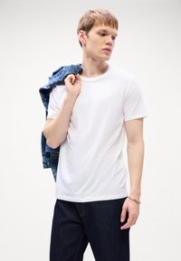 Ung mann i hvit t-skjorte og mørk jeans holder en blå cowboyjakke over skulderen, mens han ser sidelengs mot en ensfarget bakgrunn.