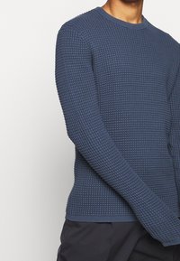 Homme portant un pull en tricot texturé bleu avec un col rond et des manches longues, associé à un pantalon noir sur un fond blanc.