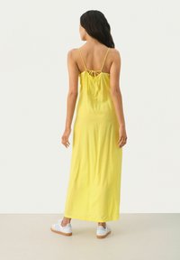 Gelbes Maxikleid mit Spaghetti-Trägern, gerafftem Rückendetail und glatter Textur, kombiniert mit weißen Sneakers und Gummisohle.