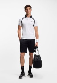 Weißes Poloshirt mit schwarzen Akzenten, Logo auf der Brust, kombiniert mit schwarzen Shorts. Inklusive schwarzen Sneakers und einer passenden Schultertasche.