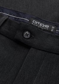 Donkergrijze broek met een denim textuur, voorzien van een zwarte knop en een label met de tekst "ombre Quintessence M/32" aan de binnenkant van de tailleband.