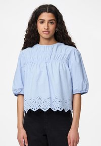 Lyseblå bomullsbluse med puffede ermer, innsamlet liv og skjært kant med broderte utklipp.