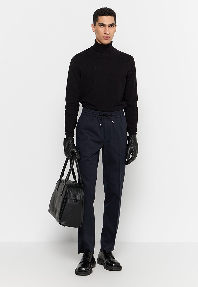 Schwarzer Rollkragenpullover, dunkelblaue Jogginghose mit Kordelzug, schwarze Handschuhe und Lederhandtasche. Das Modell steht mit neutralem Ausdruck und flachen Schuhen.