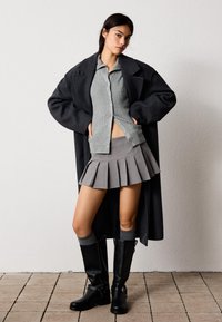 Šedý rebrovaný kardigán vrstvený pod dlhým čiernym kabátom, kombinovaný so skladanou šedou mini sukňou a čiernymi čižmami po kolená s prackami.