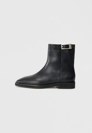 Black leather ankle boot with pointed toe, flat sole, and silver buckle strap near the top.