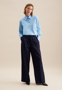Camicia azzurra chiara con colletto a punta, abbinata a pantaloni navy a vita alta e gamba larga, e décolleté nere a punta.