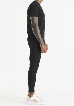 Schwarzes Baumwoll-T-Shirt, eng anliegend, mit rundem Ausschnitt. Kombiniert mit schwarzen Stretch-Leggings. Weiße Schuhe mit goldenen Akzenten. Seitenansicht.