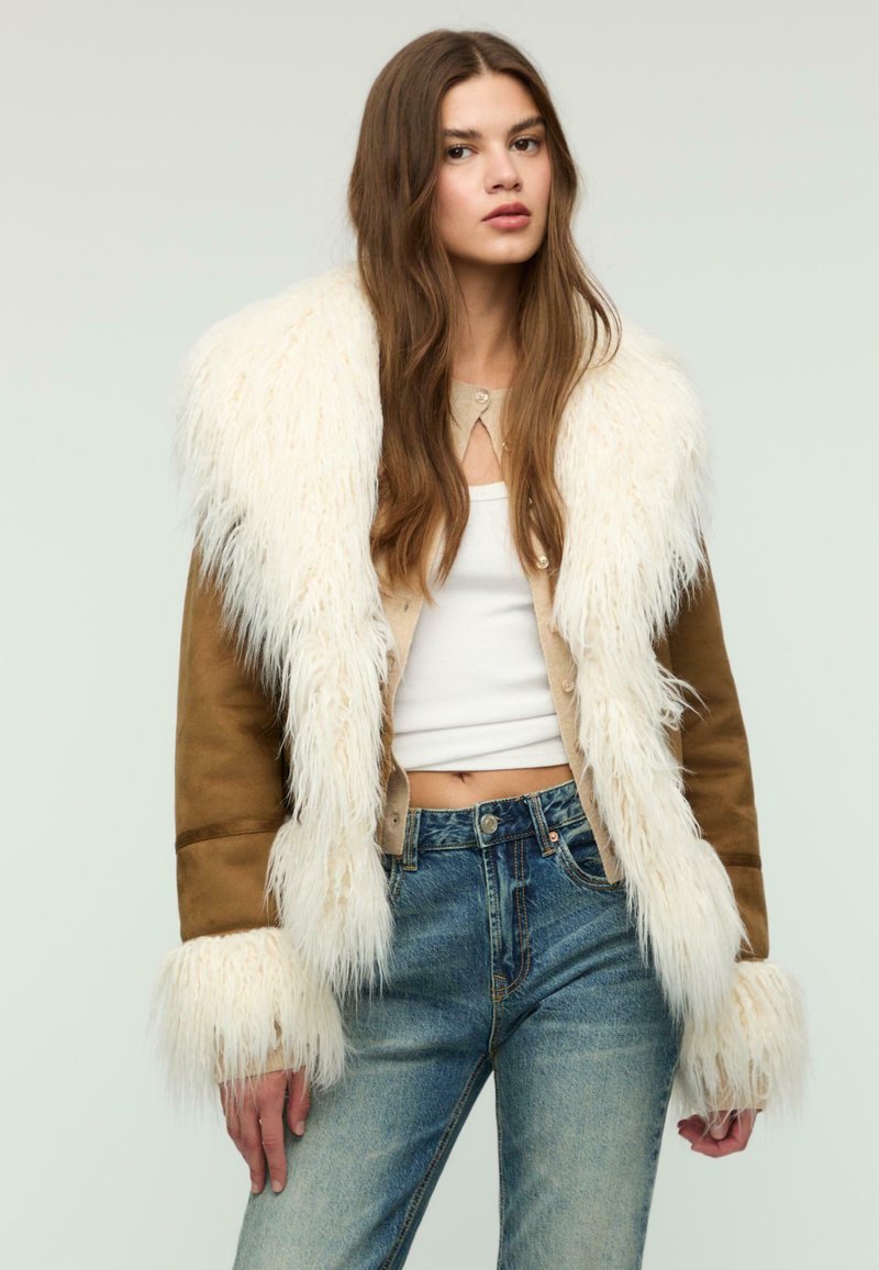 Kvinde iført en brun ruskindsjakke med hvid pelskant, en hvid crop top og blå højtaljede jeans foran en ensfarvet lys baggrund.