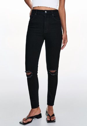 Frau trägt hoch taillierte schwarze Skinny-Jeans mit zerrissenen Knien und schwarze Sandalen mit Absatz, steht vor einem schlichten Hintergrund.
