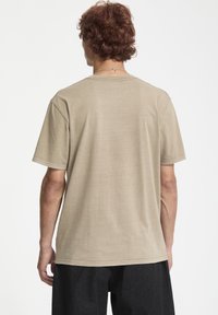 Beige kortærmet t-shirt, rund halsudskæring, blød tekstur, lige snit, ensfarvet design. Båret med mørke bukser, set fra ryggen.