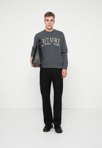 Giovane uomo che indossa un maglione grigio con "FUTURE est. 2018 PARIGI", pantaloni cargo neri, scarpe nere, che tiene una grande borsa di pelle nera su uno sfondo bianco.