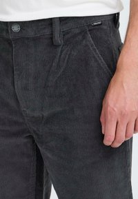 Dark grey corduroy pants with vertical ribbed texture, featuring front pockets and a button closure. Logo tag visible on waistband.