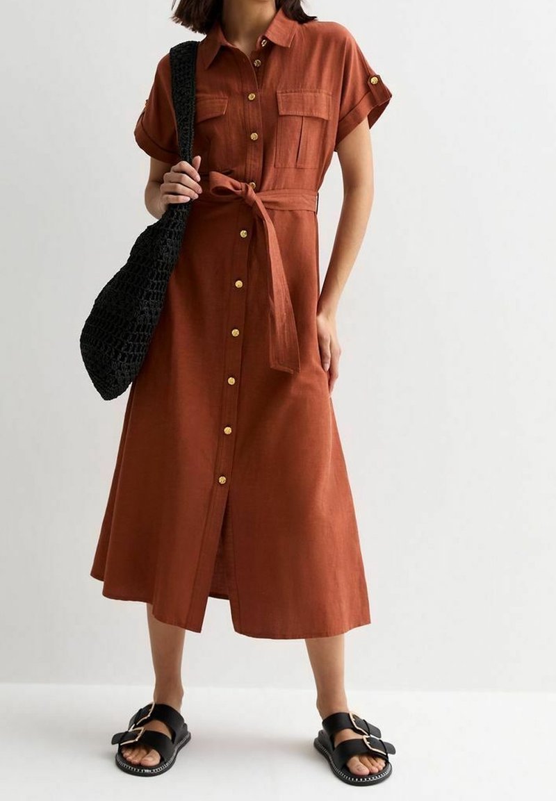 Robe à boutons de couleur rouille, longueur genou, avec des manches courtes, deux poches avant et une ceinture nouée à la taille. Associée à des sandales noires et un sac.