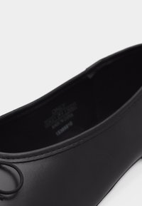 Μαύρο μπαλαρίνα παπούτσι με στρογγυλεμένη μύτη και μικρή λεπτομέρεια φιόγκο, με λεία ματ επιφάνεια και εμφανή ραφή κατά μήκος της άκρης του ανοίγματος.