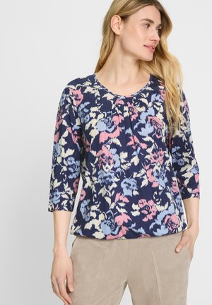 Blumenmuster Langarmoberteil in Marineblau mit pinken und hellblauen Mustern; verfügt über einen runden Ausschnitt und eine soft-texturierte Oberfläche. Kombiniert mit beigen Hosen.