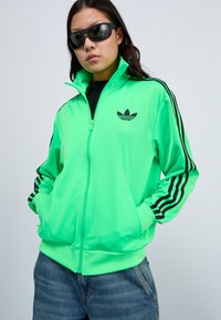 Neongrön zip-up jacka med svarta ränder på ärmarna och logotyp. Har ståkrage, framfickor och ett slätt, glänsande material.